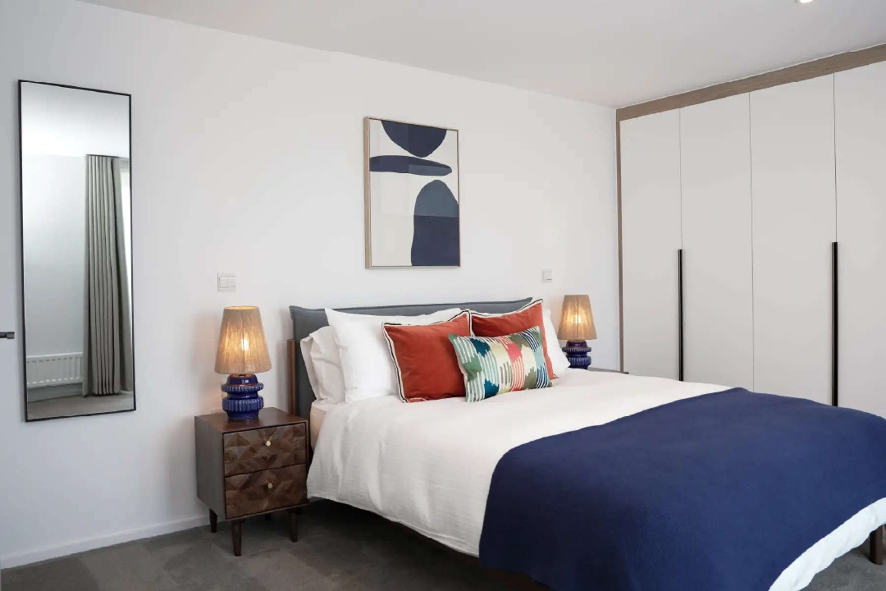 Large master bedroom, with navy double bed and accent cushions, dark wooden side tables and fitted wardrobes in a home at Brabazon, Bristol. 