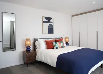 Large master bedroom, with navy double bed and accent cushions, dark wooden side tables and fitted wardrobes in a home at Brabazon, Bristol. 