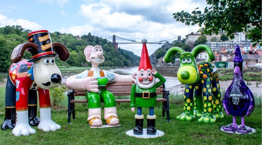 Colourful sculptures of Gromit, Wallace, Feathers McGraw and Norbot, sit on a bench by the river, with the Clifton bridge in the background.