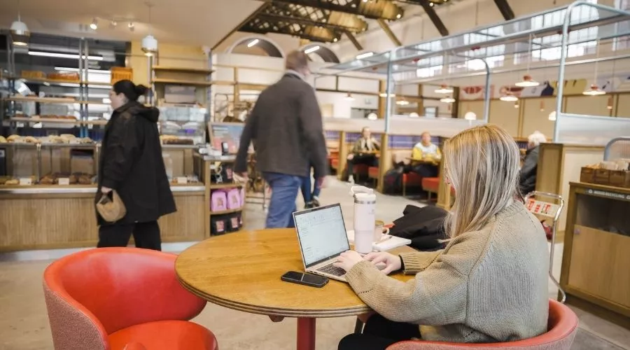A woman sits at a round table inside Mokoko Bakery at Spitfire Hangar, typing on a laptop. Other customers are visible in the background near a pastry counter. 