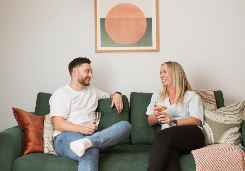Buy with a friend A man and a woman sitting on a green couch, smiling and holding drinks while engaging in conversation. The background features a framed abstract artwork with a circular design.