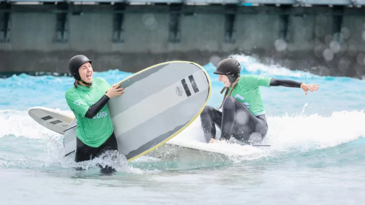 Two females laughing and smiling holding large surfboards in the shallows at The Wave surfing lagoon