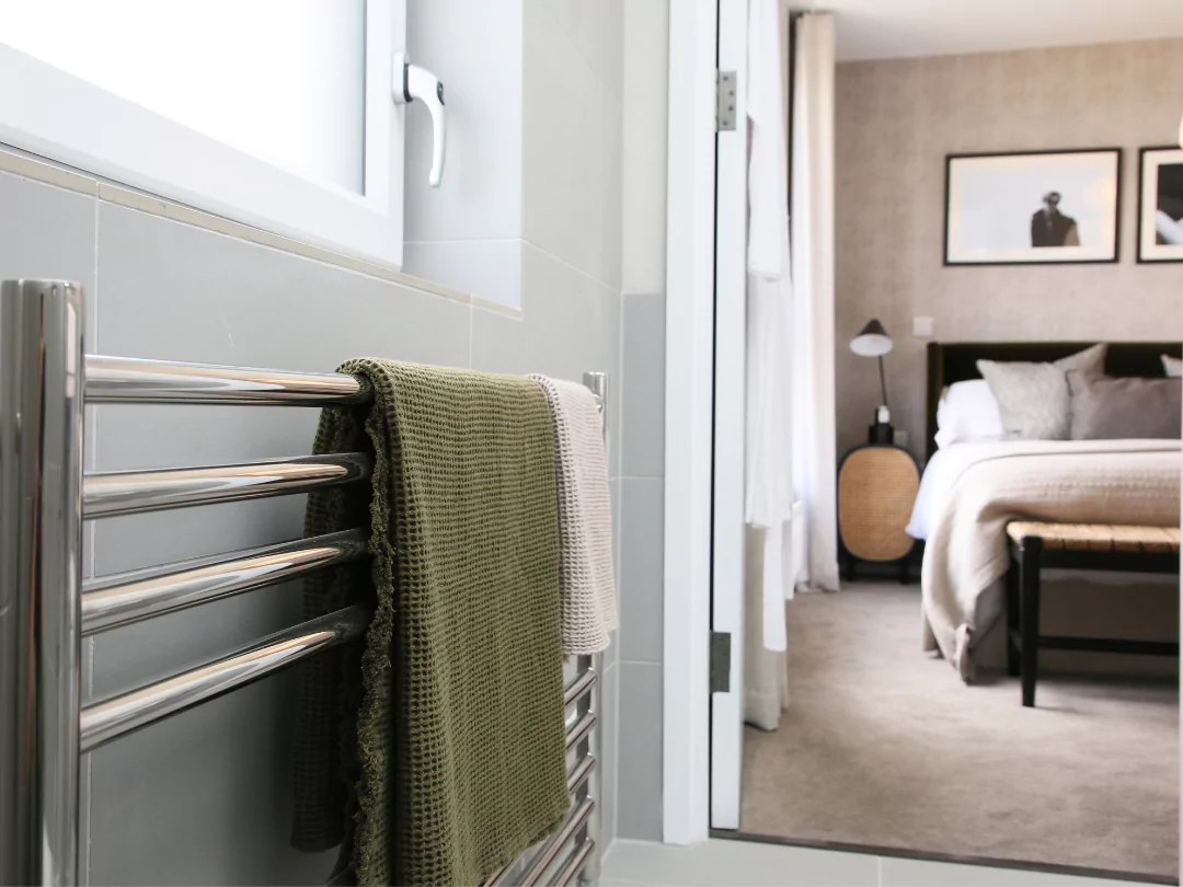 Chrome heated towel rail with dark green waffle towel hanging and view through to the primary bedroom beyond.