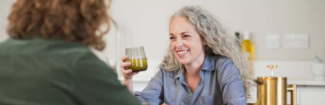 Woman with long grey hair smiling and holding a glass of orange juice, while speaking to a man, seated at a table. 