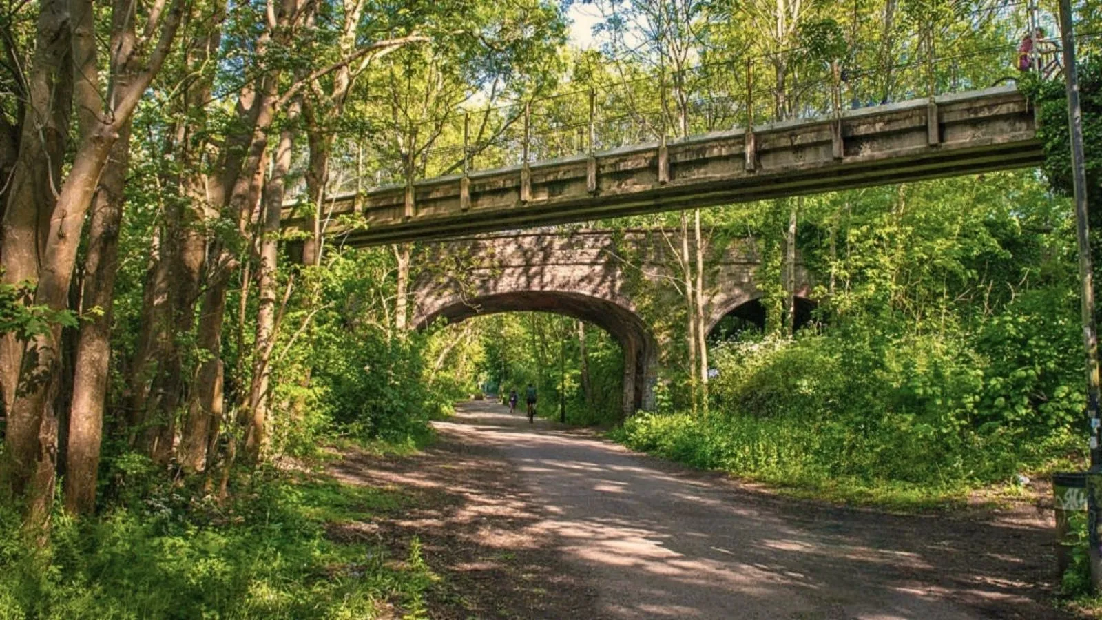 Cyclists cycling the Bristol to Bath bike path on a sunny day crossing under two bridges.