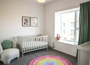 A nursery with a large window and furnished with a cot, nursing chair and colourful circular rug.
