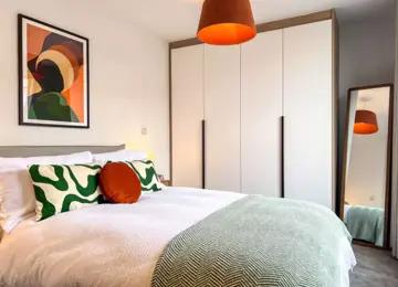 Light and modern primary bedroom, showcasing a double bed with white linen, assorted green pillows, a green throw. Behind is built in white wardrobe and a free standing full length mirror. 